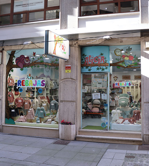 Vista exterior del comercio, dónde vemos la entrada y los dos escaparates del mismo, uno pequeño pegado a la puerta y otro más grande separado por una columna del edificio