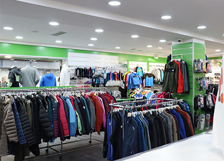 Vista del interior de la tienda, dónde podemos ver varios percheros, de pie y de pared, con múltiples prendas de ropa, tanto deportiva como de invierno. También hay percheros en una columna.