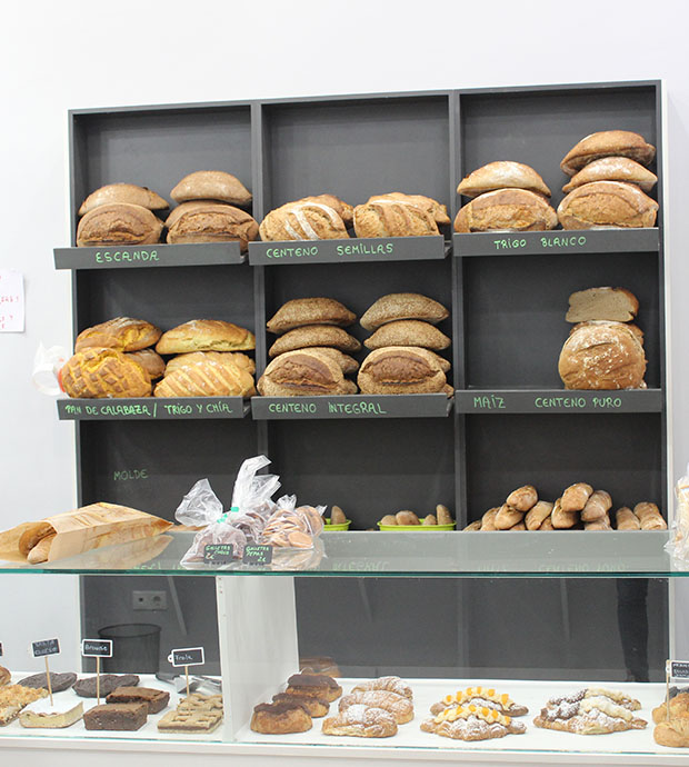 Vista del interior del local, dónde podemos ver una estantería con hogazas y barras de pan de diversos tipos. En primer plano tenemos un expositor mostrador con bollería, pasteles y tartas, sobre el hay unas bolsas de galletas y una bolsa de papel con dos barras de pan