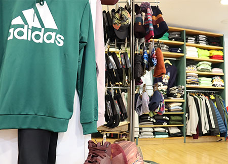 Vista del interior de la tienda, dónde podemos ver varias estanterías con ropa doblada en las baldas o en perchas, gorros y en primer plano una sudadera verde de Adidas