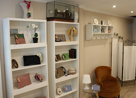 Vista del interior del comercio, dónde se pueden ver dos estanterías con bolsos de diferentes tamaños y colores, acompañados por figuras, cuadros y fotos de adorno. También vemos un un sillón y una mesita de cristal con una lámpara y otro bolso.