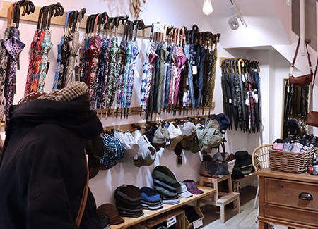 Vista del interior de la tienda dónde podemos ver paraguas colgados de percheros de pared, sobre otros percheros con gorros para la llluvia y un expositor bajo con gorras. También se ve parcialmente un expositor con otros complementos