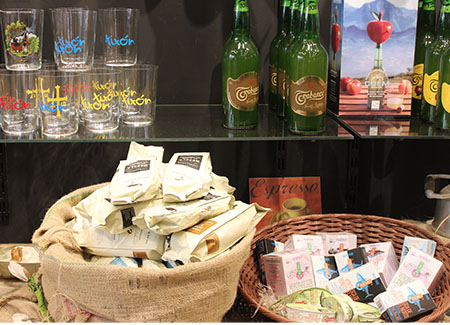 Primer plano de unas cestas de mimbre, una con paquetes de café y otra con conservas artesanales. Al fondo vemos parte de un expositor con baldas de cristal con botellas y vasos de sidra, los vasos están decorados con motivos asturianos y de Xixón